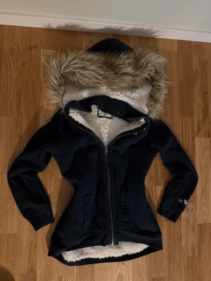 Mörkblå teddyjacka från Abercrombie - Mysig mörkblå teddyjacka från Abercrombie med fluffig fuskpäls på huvan och vit teddyfoder på insidan. Jackan har broderad logga på bröstet, dragkedja framtill och två fickor. Perfekt för kalla dagar när du vill vara både varm och snygg.