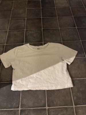 Vit croppad t-shirt från 157 XS - En enkel och fräsch vit t-shirt från 157 i croppad modell. T-shirten har rund hals och korta ärmar, tillverkad i mjuk 100% bomull. Perfekt för dig som gillar minimalistisk stil och vill ha en basic topp i garderoben.