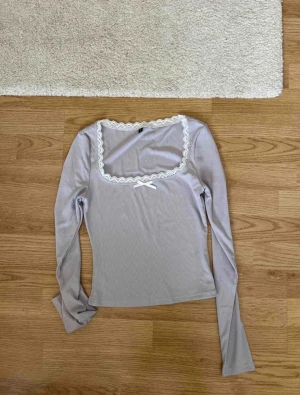 Ljus grå långärmad topp med spets - Supersöt ljus blå långärmad topp med fyrkantig ringning, dekorerad med vit spetskant och en liten rosett framtill. Toppens material är ribbad och den har en feminin och trendig vibe, perfekt att styla med jeans eller kjol.