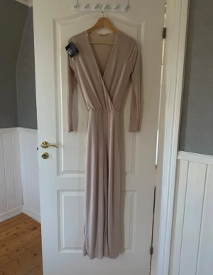 Champagnefärgad jumpsuit - Säljer en elegant jumpsuit från Nelly i champagnefärgad ton.  v-ringning och långa ärmar. Materialet är mjukt och skimrande, vilket ger en lyxig känsla. Perfekt för dig som vill sticka ut med stil.