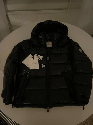  Moncler Maya - Säljer en svart  Moncler Maya med glansig finish och huva. Jackan har Moncler-logga på ärmen, dragkedja framtill och quiltad design. Perfekt för dig som vill ha en snygg och varm jacka med street-vibe. Fylld med dun och har coola detaljer som märkestaggar och justerbara muddar, NFC Scan.❄️skriv för mer frågor 🤝🏼⛷️