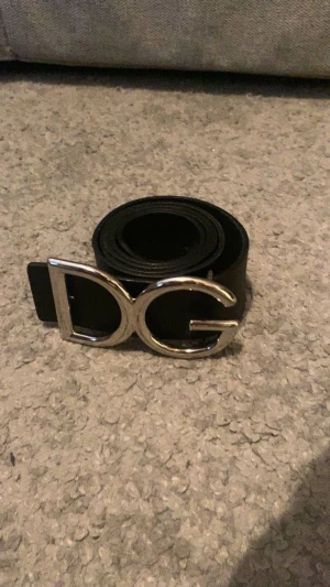 Svart DG bälte med silverdetalj - Snyggt svart bälte i läder med en stor, silverfärgad DG-logga som spänne. Perfekt accessoar för att lyfta din outfit med en lyxig touch. Klassisk design som passar till jeans eller kostymbyxor.