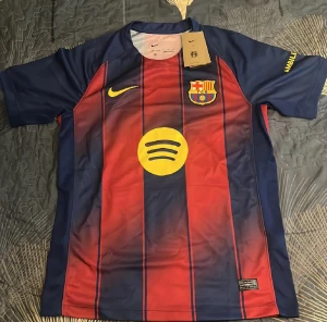 Barça Nike fotbollströja röd/blå - Säljer en officiell FC Barcelona matchtröja från Nike i röd och marinblå med gula detaljer. Tröjan har korta ärmar, vertikala ränder och klubbmärke på bröstet. Materialet är lätt polyester, perfekt för fotboll. Spotify-logga på framsidan och UNHCR-tryck på ryggen.