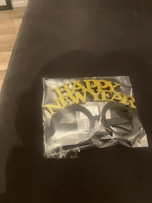Happy New Year glasögon glitter - Snygga partyglasögon med texten 'HAPPY NEW YEAR' i glittrigt guld ovanför svarta runda bågar. Perfekt accessoar för nyårsfesten, tillverkad i plast med glittrig yta på bokstäverna. Säljs i originalförpackning.