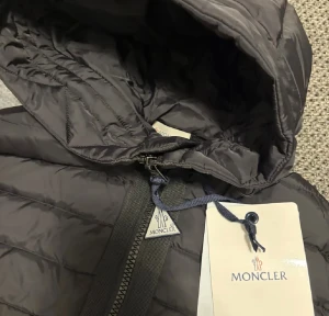 Svart Moncler pufferjacka med huva - Säljer en svart pufferjacka från Moncler med stickade ärmar och huva. Jackan har dragkedja framtill, två fickor med tryckknappar och en extra ficka med dragkedja på ärmen. Klassisk Moncler-logga på armen och ribbade muddar för extra stilpoäng. Finns alla storlekar 