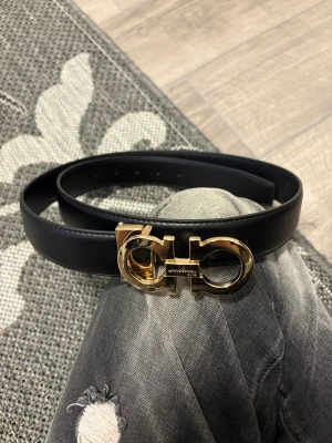 Svart skärp från Salvatore Ferragamo - Säljer ett svart bälte från Salvatore Ferragamo med en bred, stilren design och en ikonisk guldspänne med logotyp. Bältet är tillverkat i slätt skinn och passar perfekt för att lyfta din outfit med en lyxig touch.