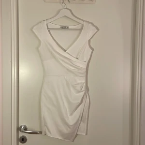 Vit omlottklänning från NLY One XS - Säljer en vit kortklänning från NLY One i storlek XS. Klänningen har omlottdesign med v-ringning och lätt rynkad detalj i sidan. Den är ärmlös och har en figurnära passform som framhäver siluetten. Perfekt för dig som gillar stilrena och enkla looks. Två små fläckar finns på ryggen!
