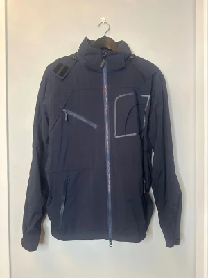 Mörkblå vindjacka från Mountain Horse - Snygg mörkblå vindjacka från Mountain Horse med huva och flera praktiska fickor med dragkedja. Jackan har sportig design, meshfoder och detaljer i grått. Perfekt för dig som gillar att vara aktiv utomhus och vill ha en jacka med cool look.