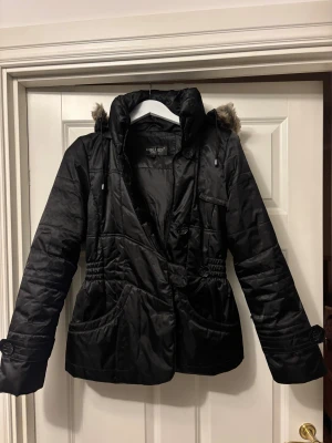 Svart pufferjacka från Hollies - Mycket bra skick! Svart pufferjacka från Hollies med snyggt quiltad design, stor avtagbar huva med fuskpäls och både dragkedja och knappar framtill. Jackan har två stora fickor och dekorativa knappar. 