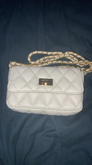 Beige axelväska med guldkedja - Snygg beige axelväska med mönster och guldfärgad kedja. Väskan har ett klassiskt lock och stängs magnetiskt i guld och ett rymligt innerfack med dragkedja. Perfekt storlek för det viktigaste och ger en lyxig känsla till din outfit.