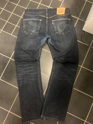 Levi's 501 mörkblå jeans straight fit - Säljer ett par klassiska Levi's 501 jeans i mörkblå tvätt med snygga slitningar. Modellen har rak passform, fem fickor och knappgylf. Tydliga kontrastsömmar och den ikoniska läderpatchen baktill. Perfekta för en chill och tidlös look.