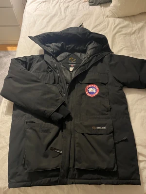 Svart dunjacka från Canada Goose M - Säljer en svart dunjacka från Canada Goose i storlek M. Jackan har flera stora fickor, huva och klassisk Canada Goose patch på bröstet. Perfekt för kalla dagar med sin tjocka och isolerande design. Stängs med dragkedja och har snygga detaljer.