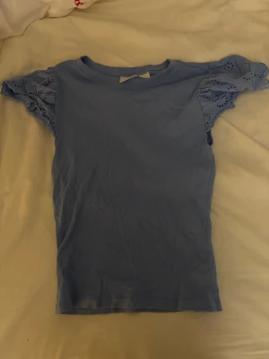 Blå blus med spetsärmar från Gina Tricot - Supersöt ljusblå blus från Gina Tricot med korta puffärmar i broderad spets. Toppen är ribbad i kroppen och har rund hals. Perfekt för dig som gillar romantiska detaljer och vill ha en feminin look. Säljer då den inte kommer till användning längre!