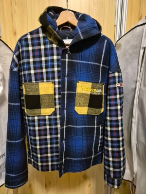  jacka Tommy Jeans - Säljer en fet rutig oversize jacka från Tommy Jeans i blå, svart och gul. Stor i storleken. Size M men som size L. Jackan har huva, två stora bröstfickor med gul-svart rutmönster och broderad logga. Materialet är mjukt och känns som ullblandning, perfekt för höst och vinter. Långärmad och med tryckknappar framtill. 