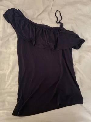 Svart offshoulder topp från Name It - Säljer en svart offshoulder topp från Name It med volang längs halsringningen och ett justerbart axelband på ena sidan. Toppens stil är riktigt snygg och passar perfekt till dig som gillar trendiga detaljer. Materialet är mjukt och skönt, troligen bomull.