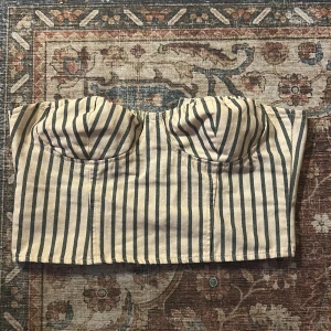 Randig korsett-topp från NA-KD - Säljer en snygg korsett-topp från NA-KD i storlek 36. Toppen är croppad och har vertikala blå ränder på beige botten, med formade kupor och dragkedja bak. Gjord i bomullsmaterial som känns mjukt mot huden. Perfekt för dig som vill ha en trendig och cool look.