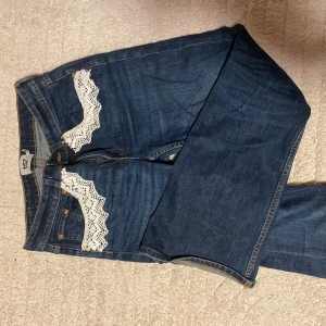 Blå jeans med spetsdetaljer - Snygga blå jeans från 157 med vita spetsdetaljer på både fram- och bakfickor. Spetsen är påsydd, har även experimenterats med byxorna på andra sätt, därav är bakfickorna lite slitna o har lite streck. Perfekt för dig som vill sticka ut med din stil.