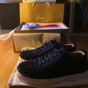 Christian Louboutin sneakers med nitar - Säljer ett par mörkblå sneakers från Christian Louboutin med coola svarta nitar på tån. Skorna har snörning, vit platt sula och är tillverkade i mocka för en lyxig känsla. Perfekt för dig som vill sticka ut med en edgy och exklusiv stil. 