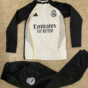Real Madrid träningströja Adidas - Snygg långärmad träningströja från Adidas för Real Madrid med svart-vita paneler, gula detaljer och klubbmärke på bröstet. Tröjan har half zip och är tillverkad i polyester, perfekt för fotbollsträning. Matchande svarta byxor med klubbmärke ingår.