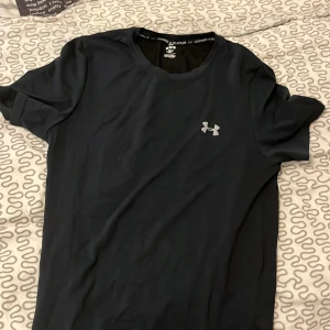 Svart Under Armour t-shirt M - Svart t-shirt från Under Armour i storlek M. Stilren design med logga på bröstet och diskret logga i nacken. Tillverkad i mjukt och lätt polyester som andas, perfekt för träning. Klassisk rund hals och korta ärmar för en sportig look.