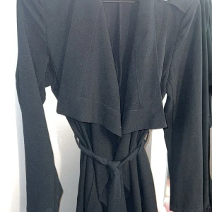 Svart jacka från Object XS - Svart jacka från Object i storlek XS. Jackan har långa ärmar, bred krage och knytband i midjan för en snygg siluett. Materialet är mjukt och har en lätt struktur. Perfekt för dig som gillar stilrena och enkla plagg med en modern touch.