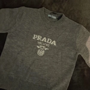 Grå stickad tröja från Prada - Grå stickad tröja från Prada med broderad logga och texten 'PRADA MILANO' framtill. Tröjan har rund halsringning, långa ärmar och breda ribbade muddar vid ärmslut och nederkant. Perfekt för dig som gillar klassisk och stilren design.