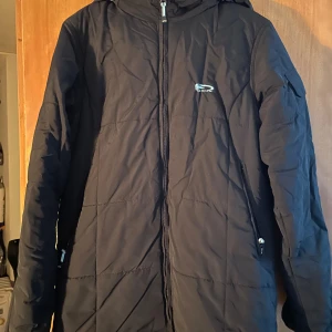 Svart lång pufferjacka från Neill - Säljer en svart pufferjacka från Neill med huva och hel dragkedja framtill. Jackan har två sidofickor med dragkedja och ett litet logomärke på bröstet. Perfekt för kalla dagar och har en stilren look som funkar till det mesta.