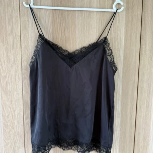 Svart linnetopp med spets från Zara - Supersnygg svart linnetopp från Zara med tunna axelband och v-ringning. Toppen har fina spetsdetaljer längs halsringning och nederkant som ger en extra touch. Perfekt att styla med jeans eller kjol för en trendig look.
