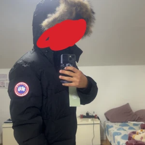 Svart dunjacka från Canada Goose - Säljer en svart dunjacka från Canada Goose med stor huva och snygg pälsdetalj. Jackan har klassisk patch på ärmen och är perfekt för kalla dagar. Fodret är mjukt och jackan har en normal passform med långa ärmar. Grym stil för vintern!