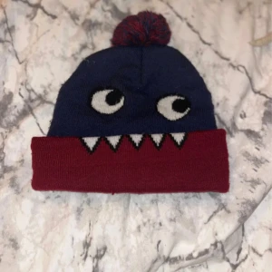 Blå och vinröd mössa med monstertryck - Säljer en cool mössa från Lindex med blå ovandel och vinröd uppvikt kant. Framsidan har ett monstertryck med ögon och vita tänder, samt en matchande blå och vinröd tofs på toppen. Perfekt för dig som gillar roliga detaljer och vill sticka ut.