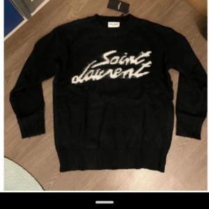 Svart stickad Saint Laurent tröja - Svart stickad tröja från Saint Laurent med vit logotyp i graffiti-stil på bröstet. Tröjan har rund hals, ribbade muddar och lång ärm. Tillverkad i Italien och har en mjuk, lyxig känsla som gör den riktigt snygg och chill.