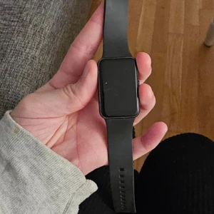 Svart smartklocka med laddare - Säljer en stilren svart smartklocka med rektangulär skärm och silikonarmband. Klockan har en modern look och kommer med tillhörande laddkabel. Perfekt för dig som vill ha koll på tiden och dina aktiviteter direkt på handleden.