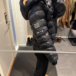 Svart pufferjacka från Peak Performance - Säljer en svart, glansig pufferjacka från Peak Performance med huva och dragkedja framtill. Jackan har en cool patch på ärmen, muddar vid ärmslut och justerbar huva. Inga defekter. Passar både tjejer och killar! Nypris ca 2000-3000.
