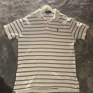 Randig pikétröja från Polo Ralph Lauren - Vit pikétröja från Polo Ralph Lauren med tunna marinblå och ljusrosa ränder. Klassisk krage, korta ärmar och broderad logga på bröstet. Tillverkad i 100% bomull för en skön och luftig känsla. Perfekt för en stilren och avslappnad look.                    Liten fleck i armen men kan nog gå bort.
