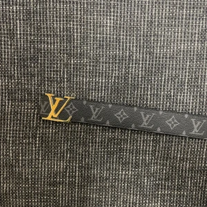 Louis Vuitton svart monogram bälte - Snyggt svart bälte från Louis Vuitton med klassiskt monogrammönster och en guldig LV-logga som spänne. Bältet är gjort i skinn och har en stilren, lyxig känsla som verkligen sticker ut. Perfekt accessoar för att lyfta din outfit.