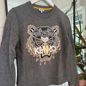 Grå Kenzo sweatshirt med tigertryck - Grå sweatshirt från Kenzo med ikonisk broderad tiger på bröstet i vitt, svart och gult. Rund halsringning och långa ärmar. Mjuk bomullskvalitet och normal passform. Perfekt för dig som vill sticka ut med ett coolt statement-plagg.