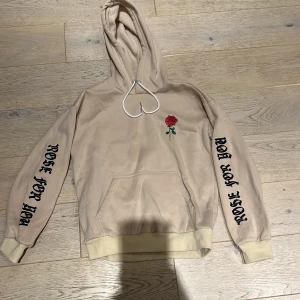 Beige hoodie med ros och tryck från MAN - Snygg beige hoodie från MAN med röd ros-tryck på bröstet och texten 'ROSE FOR YOU' i svart på båda ärmarna. Tröjan har huva med vita snören och en stor magficka. Perfekt för dig som gillar streetstyle och unika detaljer. Storlek S barn storlek