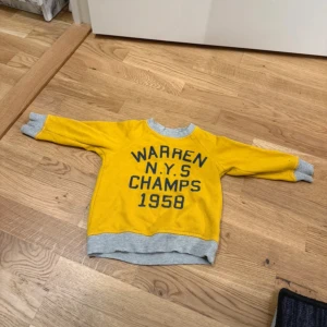 Gul sweatshirt från H&M L.O.G.G. 9-12 mån - Gul sweatshirt från H&M L.O.G.G. med tryck 'WARREN N.Y.S CHAMPS 1958' i mörkblått. Tröjan har grå ribbade muddar vid hals, ärmslut och nederkant. Perfekt för en sportig och retro look till de minsta, förekommer några fläckar samt är lite nopprig.
