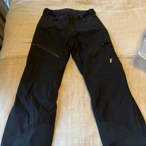 Skidbyxor Peak Performance Goretex - Svarta täckbyxor från Peak Performance med Goretex-material och RECCO-reflektor. Byxorna har justerbar midja med kardborre, dragkedja på benet och flera praktiska fickor. Perfekta för vinterns äventyr och outdoor.