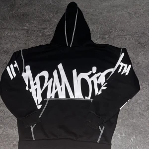 Svart hoodie med vit graffiti-text - Cool svart hoodie med stor vit graffiti-inspirerad text över bröstet och ärmarna. Tröjan har huva med kontrastsömmar och klassisk känguruficka framtill. Perfekt för dig som gillar streetwear och vill sticka ut.