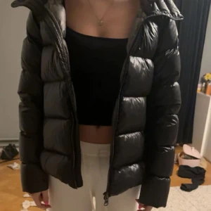 Svart Colmar Jacka - Säljer en svart pufferjacka med huva och dragkedja framtill. Jackan har en snygg, quiltad look med breda kanaler och två sidofickor. Perfekt för kalla dagar och har en modern, loose passform som är enkel att styla till olika outfits.