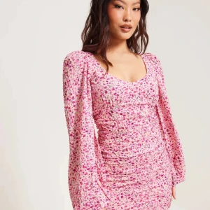 Rosa blommig kortklänning med puffärm - Supersöt kortklänning i rosa med blommigt mönster och puffiga långärmar. Klänningen har rynkade detaljer, öppen rygg med knytning och dragkedja bak. 