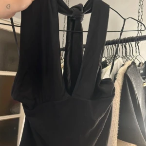 Svart omlotttopp med knyt - Svart omlottklänning från GinaTricot i mjukt material med snygg knytning i nacken. toppen har ärmlös design och djup v-ringning framtill, vilket ger en cool och modern look. Perfekt för dig som gillar minimalistisk stil och vill ha något enkelt men ändå trendigt.