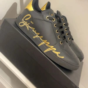 Giuseppe Zanotti - Svarta sneakers i skinn från Giuseppe Zanotti med stora guldfärgade loggor på sidorna och gulddetaljer på häl och snörhål. Klassisk rund tå och platt sula. Snygg kontrast mellan svart och guld som ger en lyxig känsla.