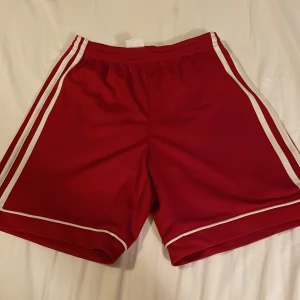 Röda Adidas shorts med vita ränder - Snygga röda Adidas shorts med vita ränder längs sidorna och broderad logga. Tillverkade i lätt och snabbtorkande Climalite-polyester, perfekta för sport eller träning. Klassisk sportig look med elastisk midja och bekväm passform.