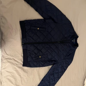 Mörkblå quiltad jacka Polo Ralph Lauren - Snygg mörkblå quiltad jacka från Polo Ralph Lauren med dragkedja framtill, två fickor med knappar och broderad logga på bröstet. Jackan har ribbade muddar och klassisk bomberjacke-look. Perfekt för dig som vill ha en stilren och sportig vibe.