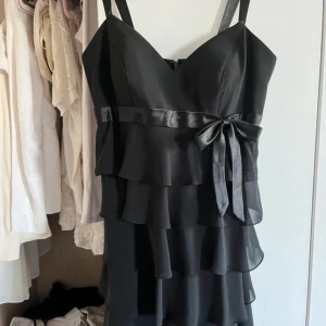 Svart volangklänning med rosett - Snygg svart kortklänning med flera lager volanger och ett satinband med rosett i midjan. Klänningen har axelband och en feminin v-ringad urringning. Perfekt för dig som gillar en chic och elegant stil med extra detaljer.