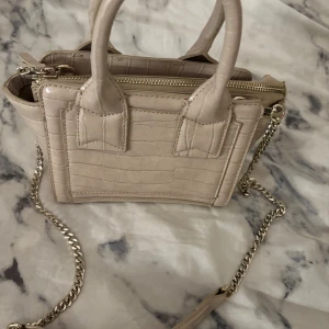 Beige handväska - Snygg beige handväska med krokodil mönster och guldfärgad axelremskedja. Väskan har två handtag och dragkedja upptill. Perfekt storlek för det viktigaste och ger en cool vibe till din outfit.