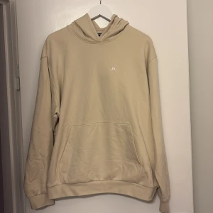 Beige hoodie från J.Lindeberg, L - Snygg beige hoodie från J.Lindeberg i storlek L. Tröjan har en klassisk huva, stor magficka och diskret vitt logotryck på bröstet. Mjuk och skön bomullskvalitet, perfekt för chill dagar. Enkel och stilren design som funkar till många outfits.