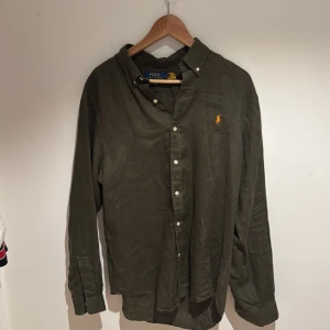 Olivgrön skjorta från Polo Ralph Lauren - Snygg olivgrön skjorta från Polo Ralph Lauren med klassisk krage och knappar framtill. Skjortan har lång ärm och den ikoniska broderade loggan i orange på bröstet. Perfekt för dig som gillar stilrena och tidlösa plagg.